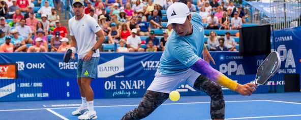 Ben Johns wearing blue shirt, white shorts, purple and yellow tie dye arm sleeves, black and gray leggings, and white hat at the 2023 Cincinnati Open.