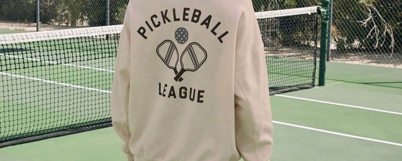 Cream sweatshirt with "pickleball league" on the back with a retro pickleball paddle design. Model wearing sweatshirt on a court.