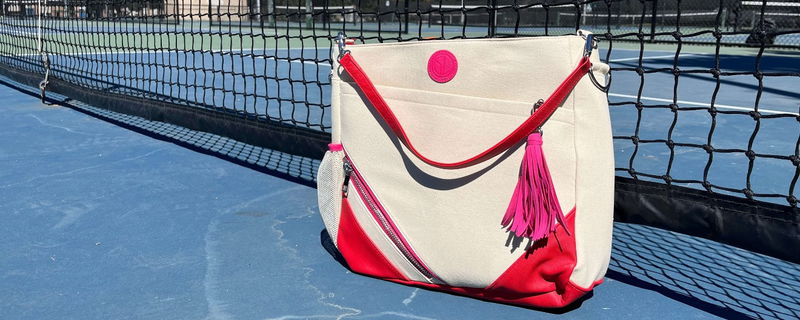 cream pickleball bag with red strap, red bottom, and pink accents sitting against a pickleball net