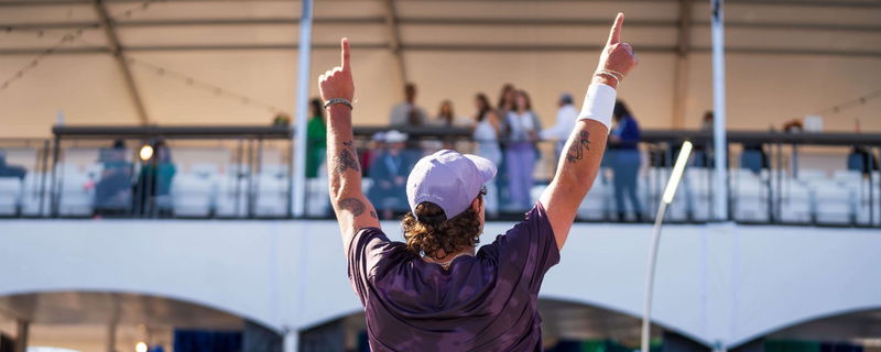 Michael Loyd turned around in a purple tie dye shirt and purple hat pointing up to the sky