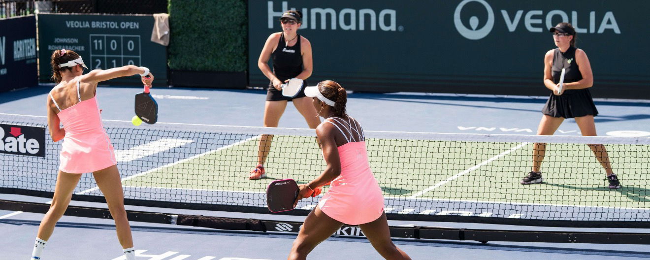 One team in matching pink dresses playing against another women's doubles team in all black.