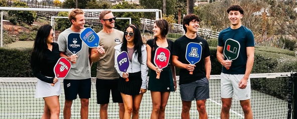 Group of people holding various college branded pickleball paddles.