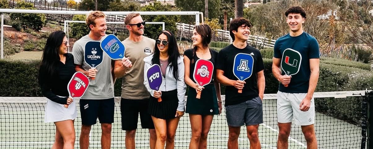 Group of people holding various college branded pickleball paddles.